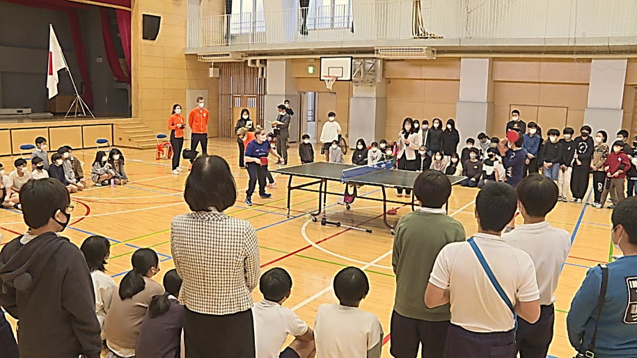 代表児童と卓球対決をするケリー選手と卓球台を囲んで応援する子どもたち