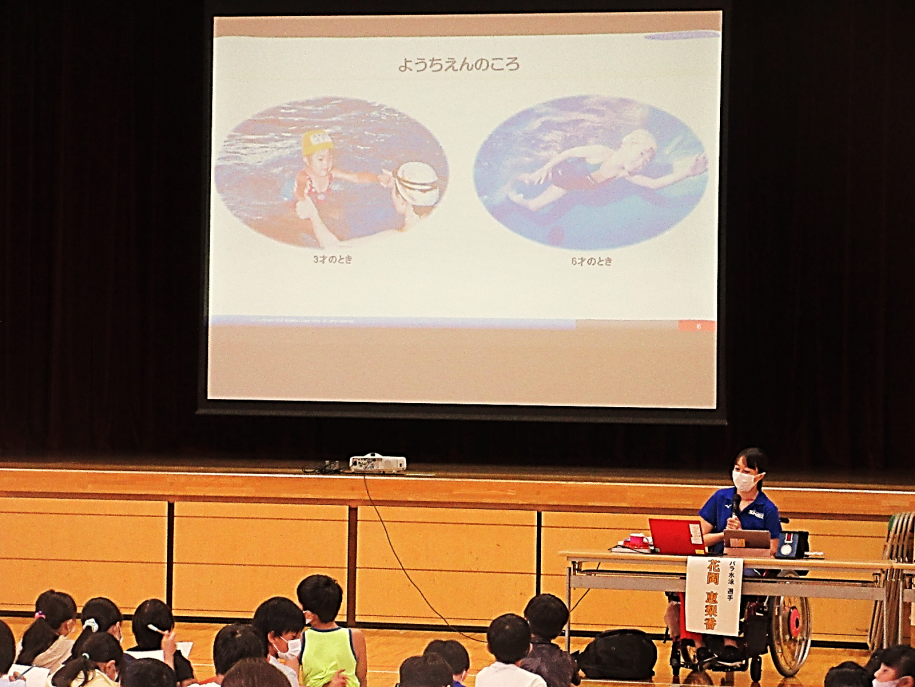 体育館のスクリーンにスライドを投影しながら小学生の前で講演するパラアスリート